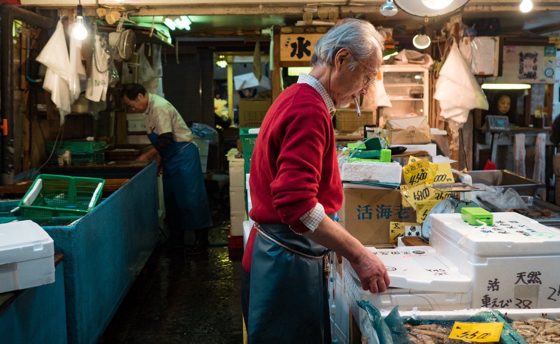 An older man in a red sweater working at a market counter.