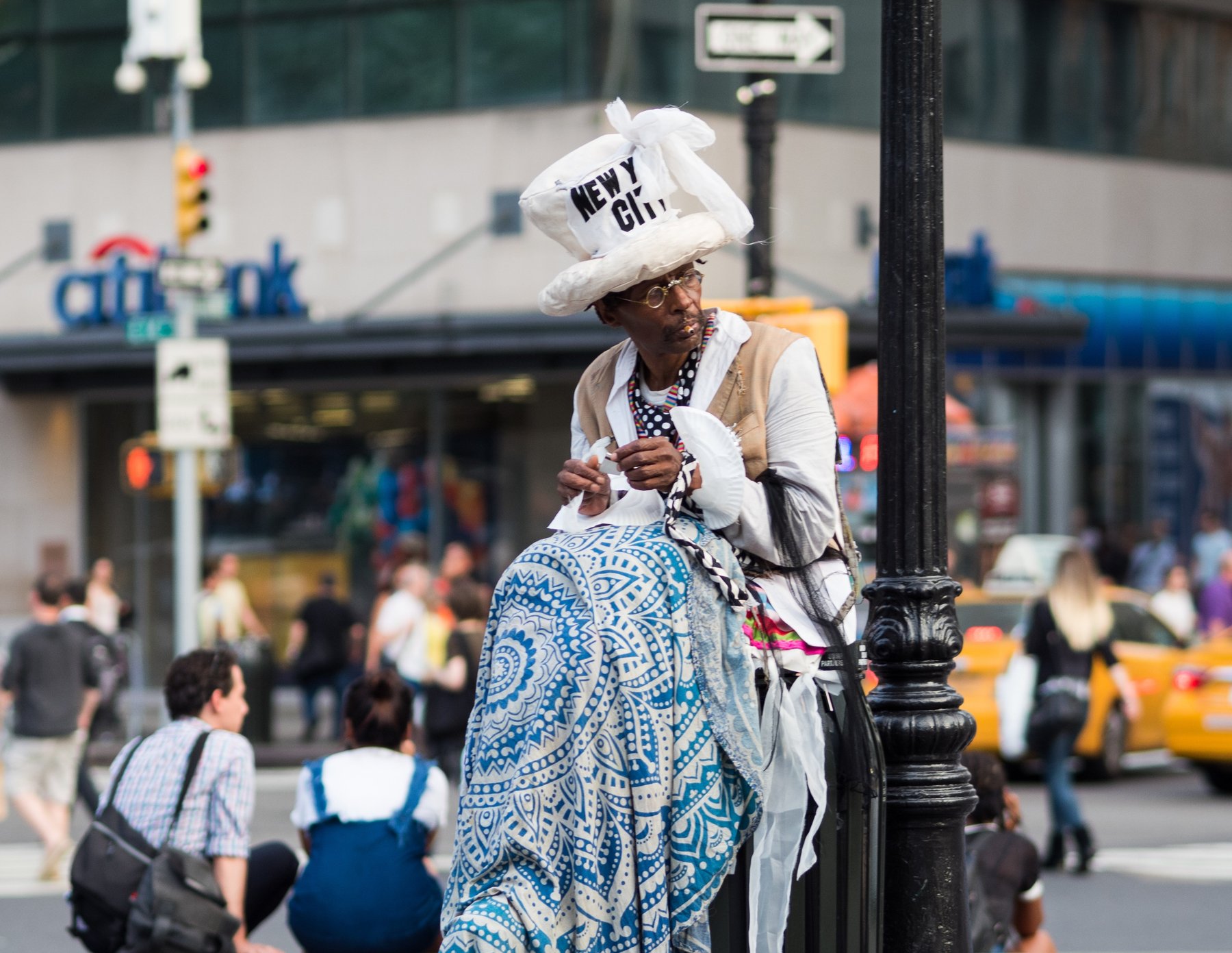 A street performer in a white headdress and patterned blue dress beside a lamppost.