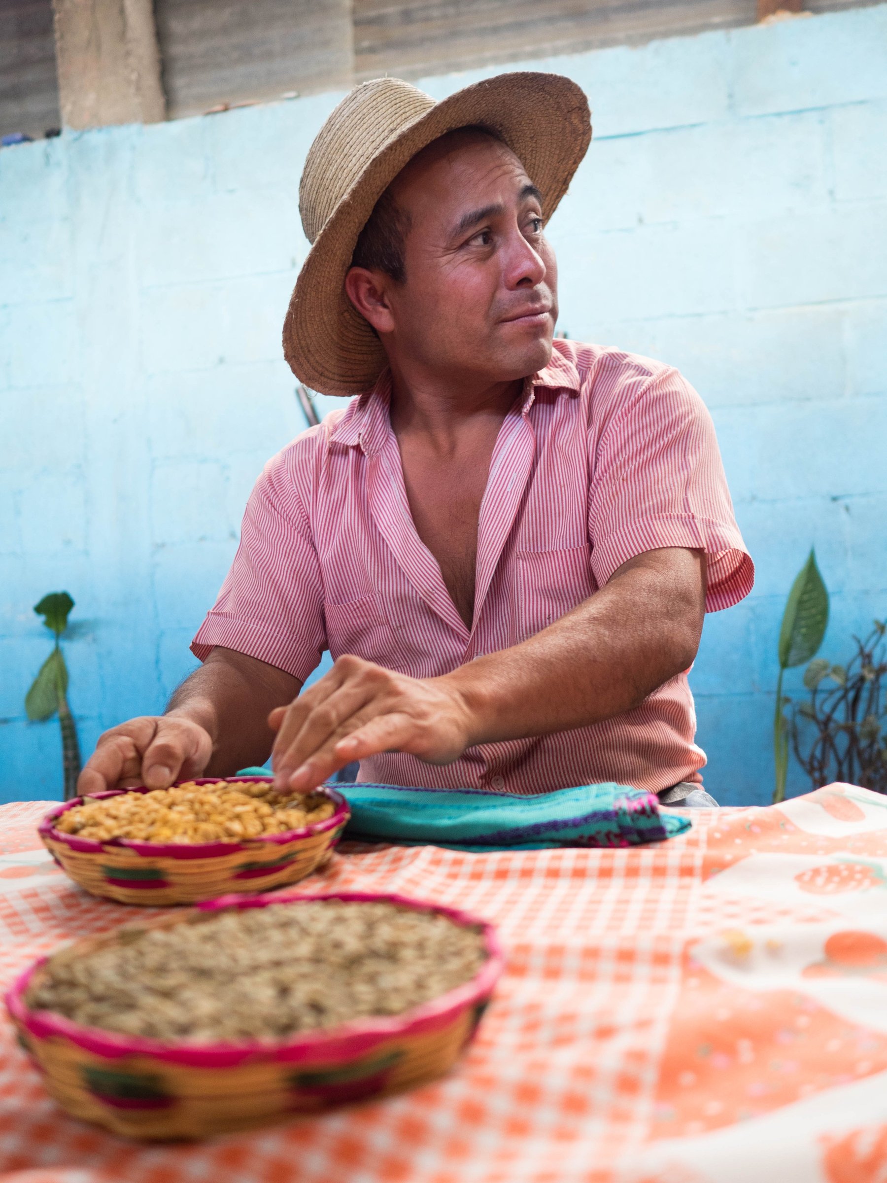 A market vendor in a straw hat and pink shirt behind a market table against a blue wall.